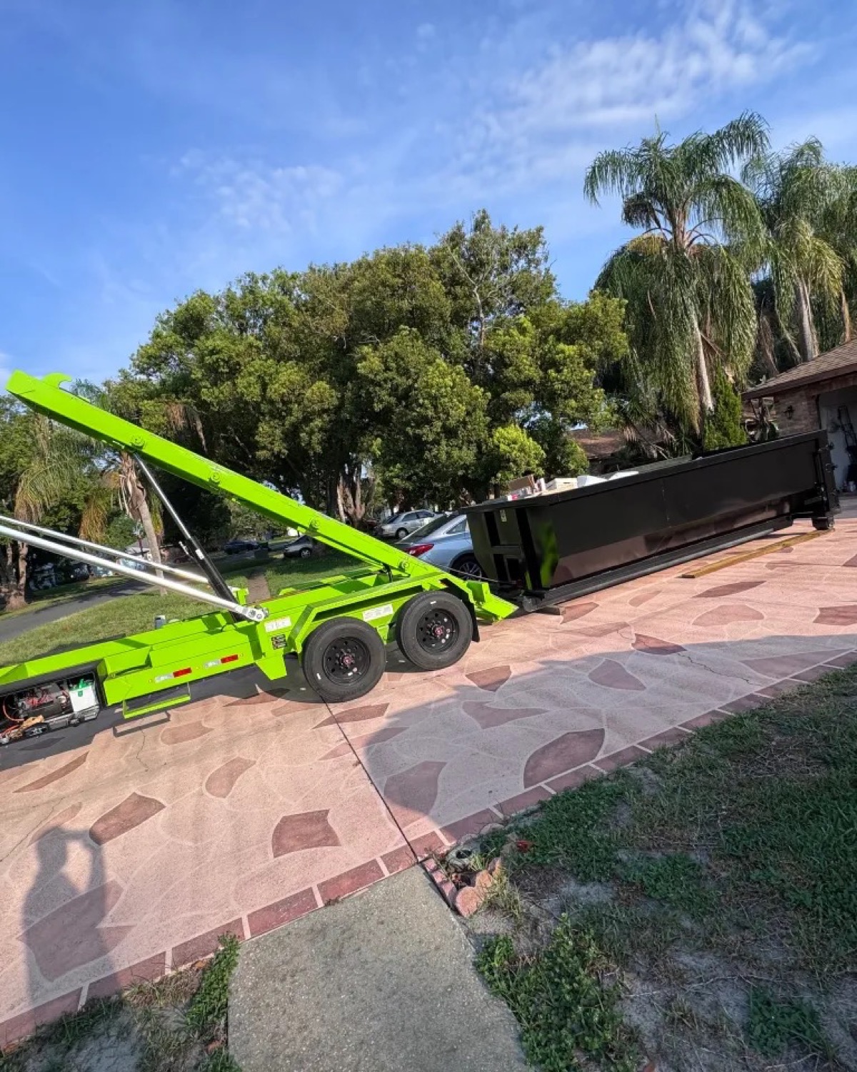 DJ's Dumpsters roll-off trailer backing a black container onto a residential driveway